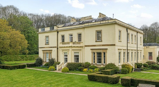 Widworthy Court exterior — a large cream-rendered two-storey house with sash windows, a balustraded balcony, and multiple chimneys, set within manicured gardens